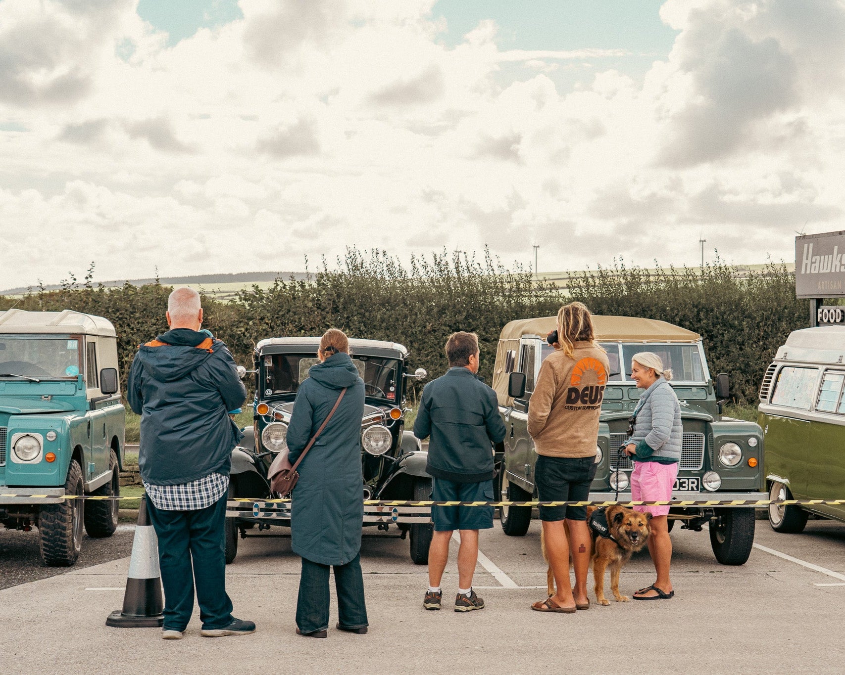 Group of people standing near vintage vans in a parking lot with a cloudy sky.